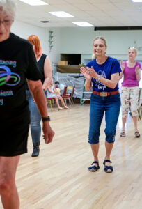 A woman in a blue shirt and jean capris claps in a line dance formation while watching other dancers.