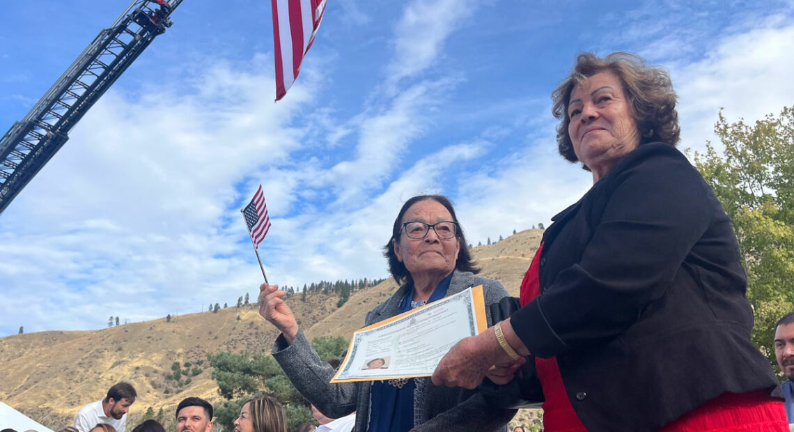 A woman on the left holds a small, American flag. She waves it. Another woman, to her right, presents her with a certificate.