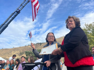 A woman on the left holds a small, American flag. She waves it. Another woman, to her right, presents her with a certificate.