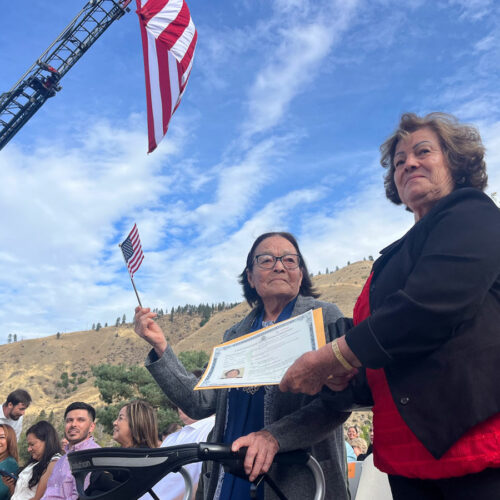 A woman on the left holds a small, American flag. She waves it. Another woman, to her right, presents her with a certificate.