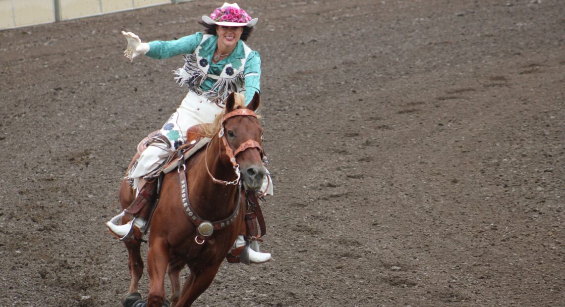 A woman wearing a flowered cowboy hat rides a dark brown horse around an arena. She waves to the crowd.