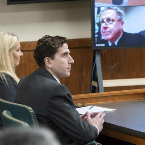 A woman and a man sit at a black table in a courtroom. A TV screen, with the judge pictured, is to the left of them.