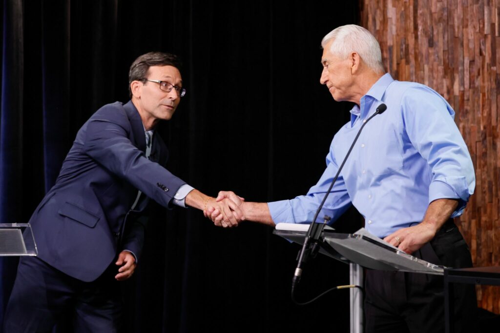 A man in a navy blue suit on shakes hands with a man in a light blue button-up shirt. He is standing behind a podium.