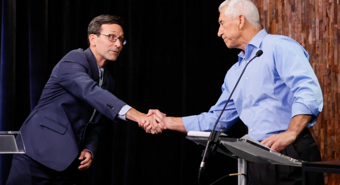 A man in a navy blue suit on shakes hands with a man in a light blue button-up shirt. He is standing behind a podium.