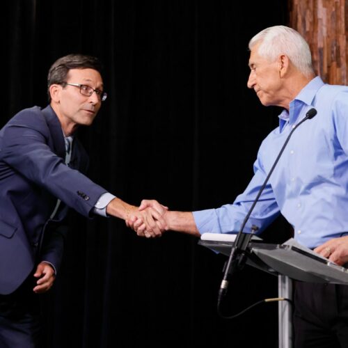 A man in a navy blue suit on shakes hands with a man in a light blue button-up shirt. He is standing behind a podium.