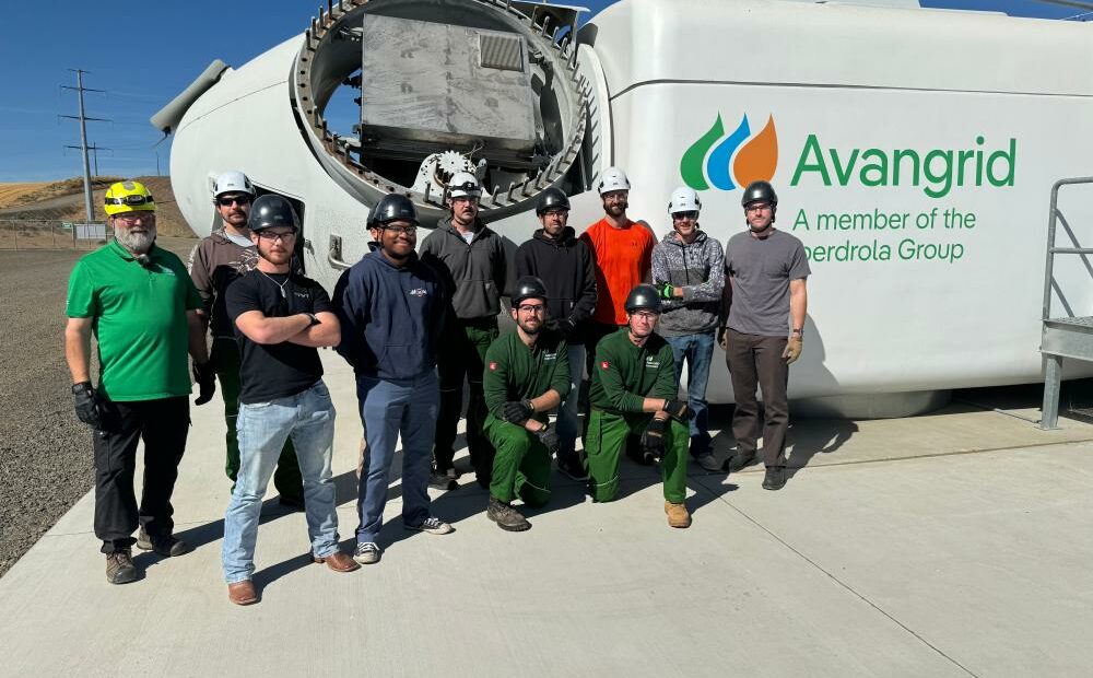 Ten men wearing hard hats stand in front of the top part of a turbine that is lying on its side on the ground. There is a sign on the equipment that says "Avangrid." They are standing on cement. The sky is blue in the background.