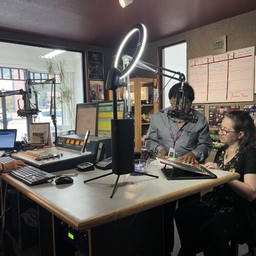 Two men and a woman gather in a sound booth in a radio station.