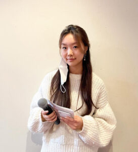 A young woman with long black hair is holding a microphone and a cellphone. She is wearing a white sweater and standing against a white background.