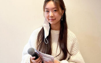 A young woman with long black hair is holding a microphone and a cellphone. She is wearing a white sweater and standing against a white background.