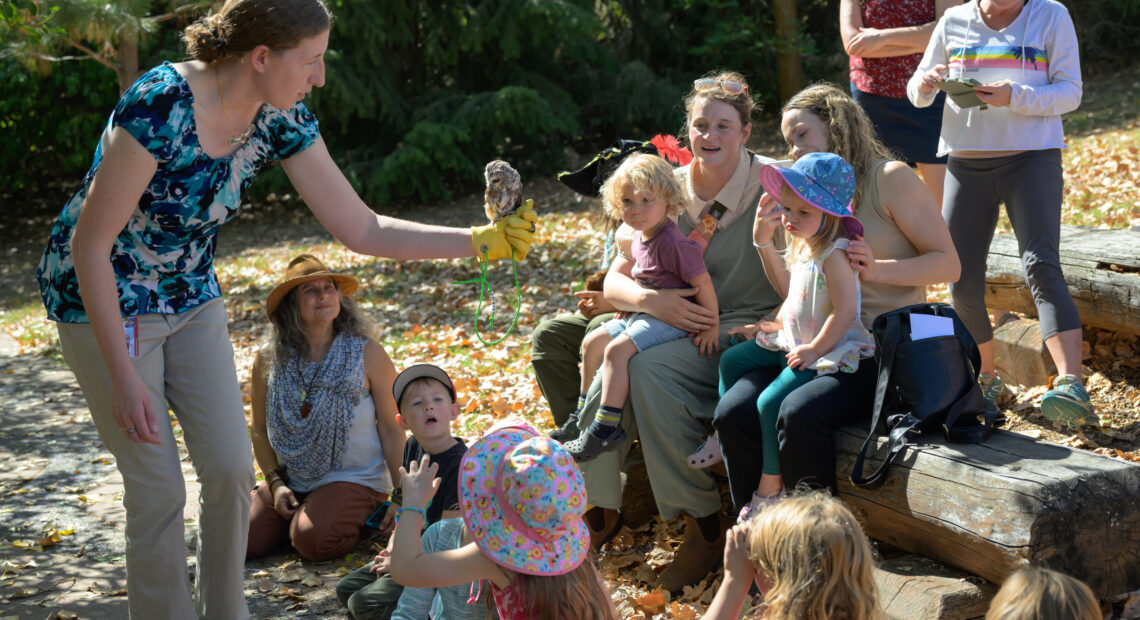 A woman wearing a blue shirt holds a small owl on her forearm. A group of children and their families are gathered around her, looking at the owl.