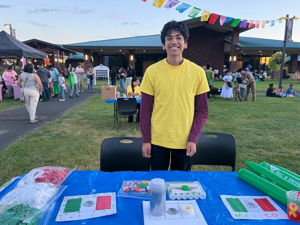 A high school student in a bright yellow t-shirt stands behind a table with a craft depicting the Mexican flag.