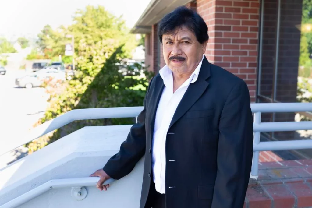 A man with a black mustache wearing a black suit poses for a portrait in front of a brick building.