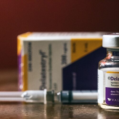 A vial of testosterone enanthate, with a needle behind it, sits on a table.