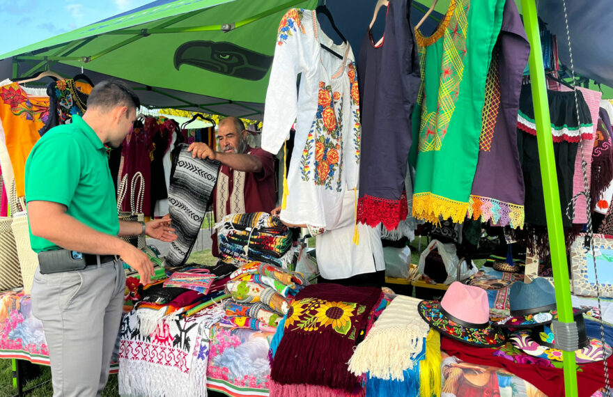 A man is standing behind a table with traditional Mexican clothing. Another man is standing in front of the table. He's looking at the items.