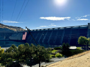 Trees and brown grass are in the foreground of the picture. Grand Coulee Dam stretches across the picture. There is an American flag hanging in front of the dam. The sky is blue.