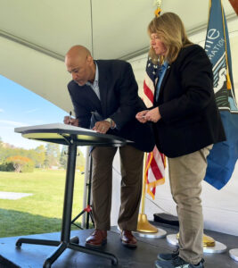 A man in a suit and a woman in a black jacket and khaki pants stand in front of a podium. The man is leaning down with a pen in his hand. Behind them are an American flag and a Bureau of Reclamation flag.