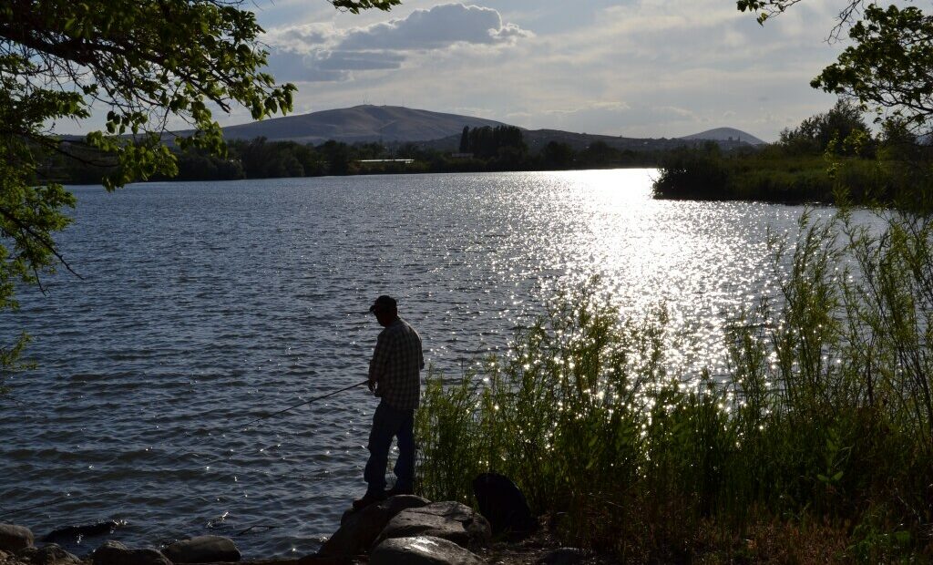 A man is silhouetted holding a fishing pole. There is water in front of him and tall grass behind him. A hill is in the distance. The sun is refelcted on the water.