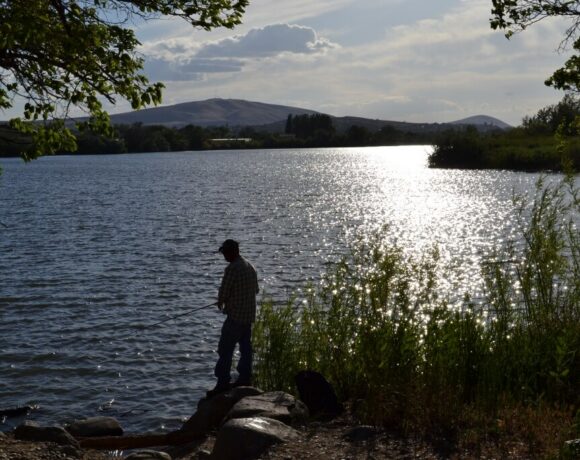 A man is silhouetted holding a fishing pole. There is water in front of him and tall grass behind him. A hill is in the distance. The sun is refelcted on the water.