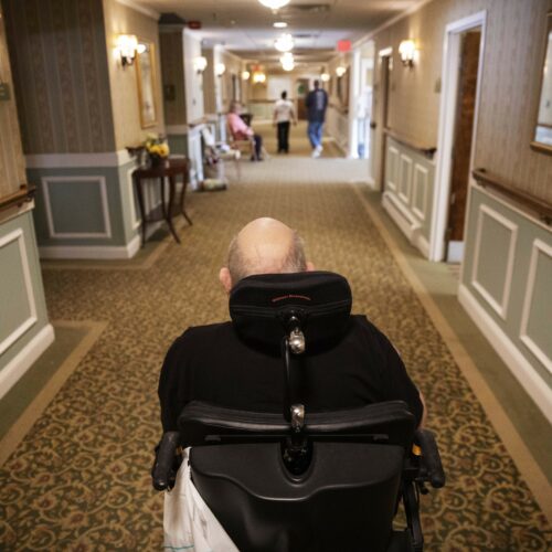 Elderly man in wheelchair in a nursing home hallway.