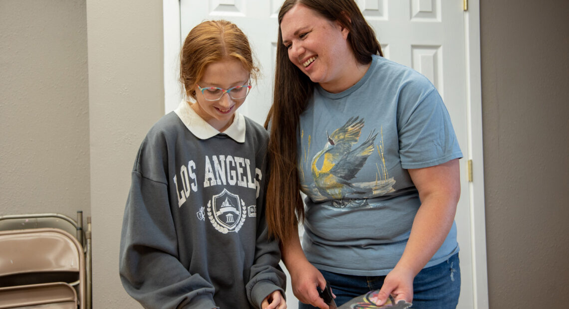 A young girl in a gray sweatshirt with a white collar stands next to a woman in a blue shirt. The girl has red hair. The woman has brown hair. They are standing behind a plastic table and in front of a white door. The woman is smiling at the girl.