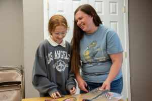 A young girl in a gray sweatshirt with a white collar stands next to a woman in a blue shirt. The girl has red hair. The woman has brown hair. They are standing behind a plastic table and in front of a white door. The woman is smiling at the girl.