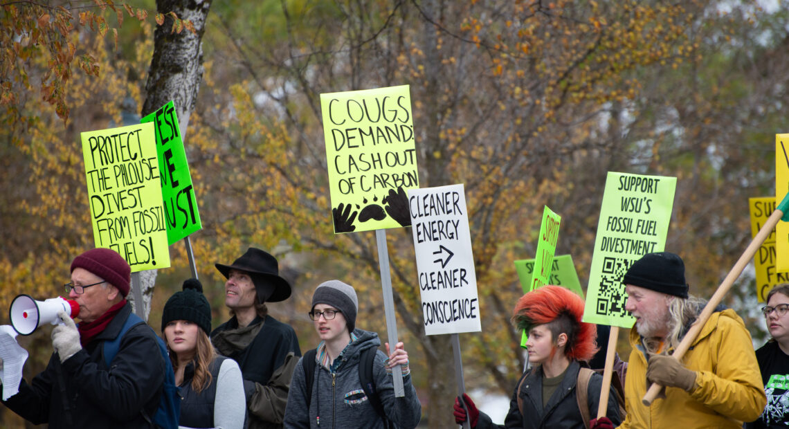 A group of people are walking, carrying brightly colored signs. Some of them read "Cougs demand cash out of carbon" and "Support WSU's fossil fuel divestment."