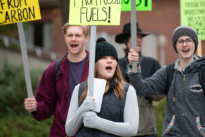 A student in a gray long sleeved shirt, black puffer vest and black beanie holds a sign at a protest. Two students stand behind her and hold signs as well.