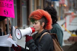 A student with a coral colored mohawk speaks into a white megaphone during a protest.