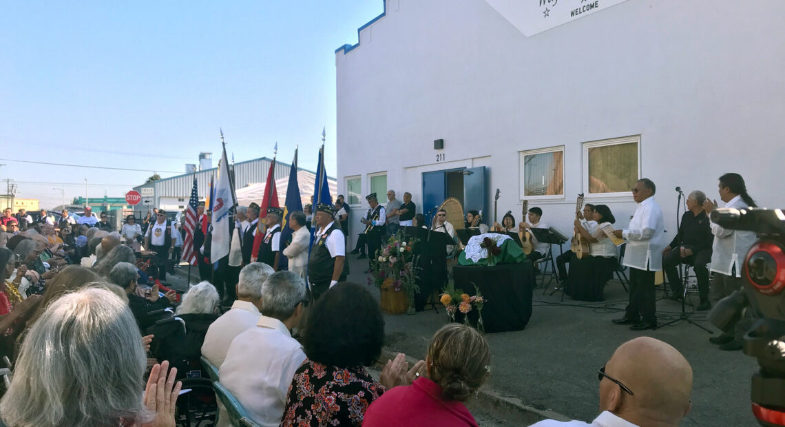 People are on a stage outside of the Filipino Community Hall of the Yakima Valley. In front of the stage, people are seated.