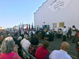 People are on a stage outside of the Filipino Community Hall of the Yakima Valley. In front of the stage, people are seated.