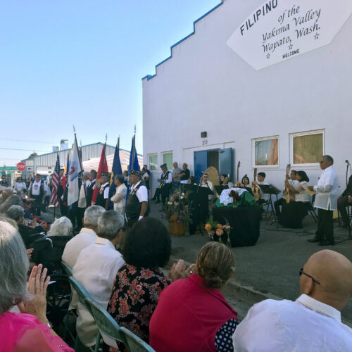 People are on a stage outside of the Filipino Community Hall of the Yakima Valley. In front of the stage, people are seated.