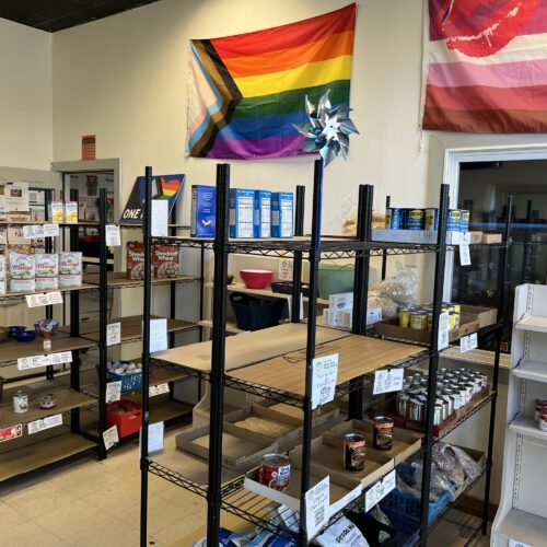 Several racks of canned goods are pictured in a room. Above the racks is a pride flag.