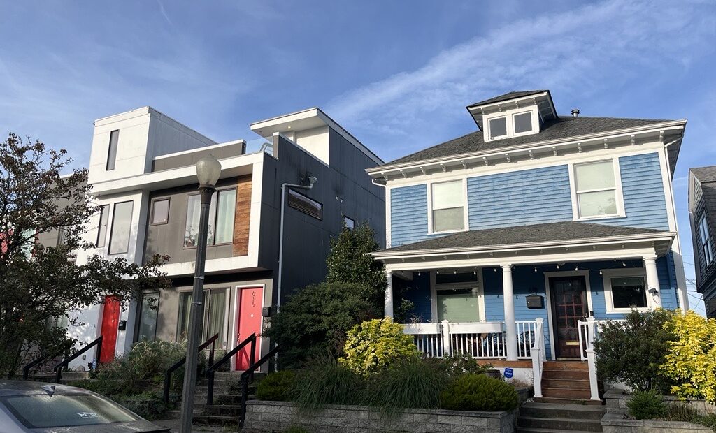 An exterior picture of two homes. The home on the left is a gray, modern style house. The home on the left is a blue Craftsman style house.