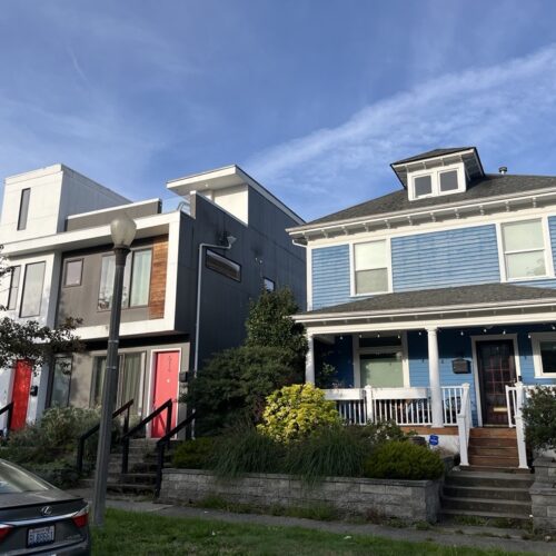 An exterior picture of two homes. The home on the left is a gray, modern style house. The home on the left is a blue Craftsman style house.