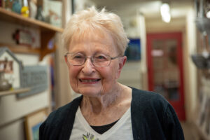 A woman with short gray hair and glasses. She's wearing a black sweater and white shirt. She's standing in a hallway that's lined with art. The art is blurry in the background.