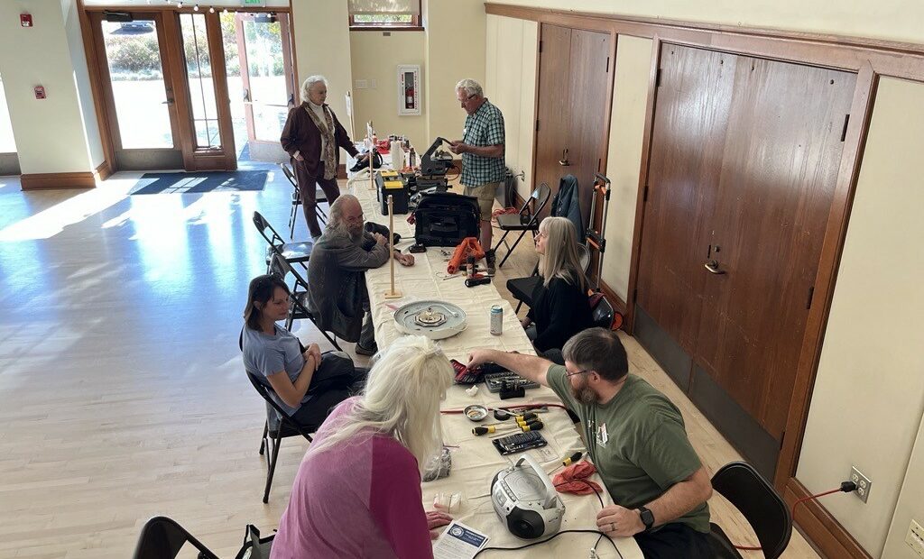 A group of people sit at two long, white tables. They are fixing a variety of items in front of them.