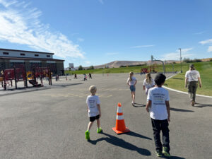 Four kids in white T-shirts that say "where kids create a greener, cooler tomorrow" on the back walk around a playground. The ground is mostly cement. There is some grass in the far right and back of the photo. Two of the kids are walking around an orange street cone.