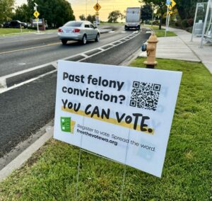 A white sign is staked in green grass next to a gray road with a silver car and white truck. There's a yellow fire hydrant in the back ground. The sign reads, "Past felony conviction? YOU CAN VOTE." There is a QR code on the sign.