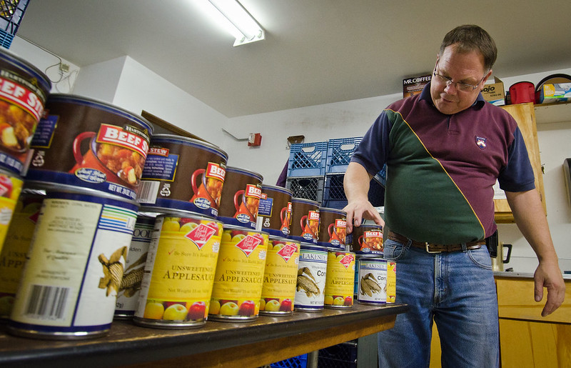 A man in a red and green shirt with blue jeans is standing next to two rows of canned food that's sitting on a table. He is in a small white room with fluorescent lights above him.