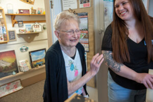 A women in a black jacket and white shirt is standing next to another woman with a black shirt and blue jeans. They are standing in front of a shelf of art.