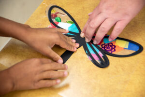 Two child hands and on adult hand are holding down a black paper dragonfly on top of a tan table. The dragonfly's wings are colored in with a bright, rainboow pf colored pieces of tissue paper and sequins. 