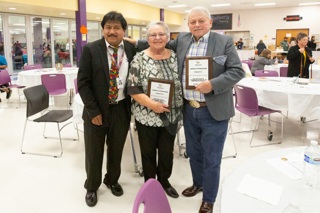 Two men and a woman pose for a photo. The woman and the man at the right of the group are holding awards.