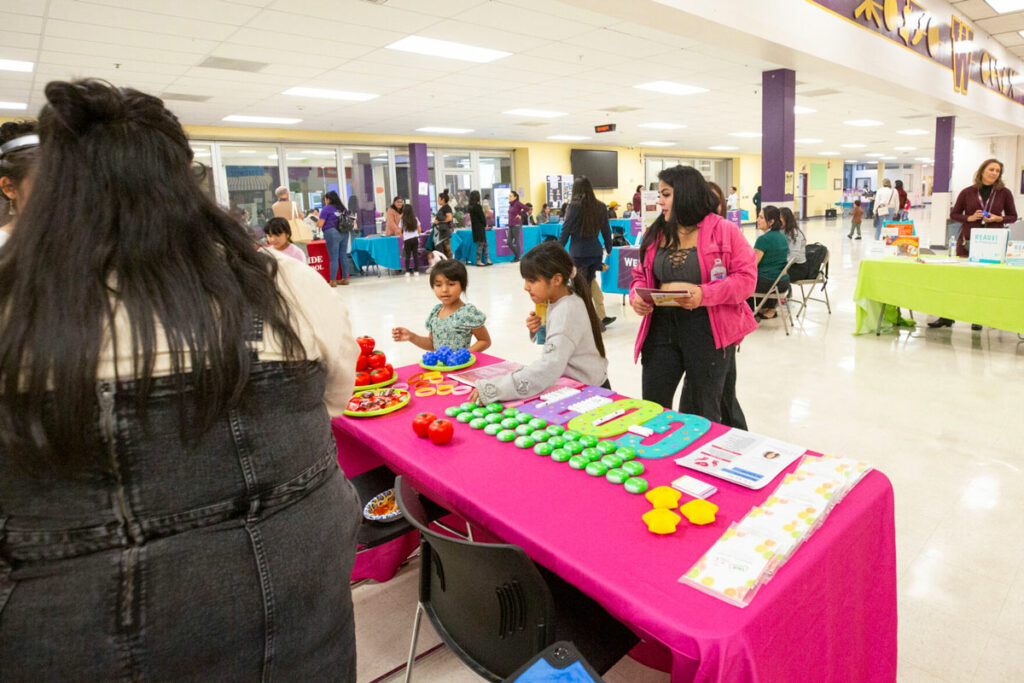 Two girls pick up items from a table. The table has a hot pink table cloth.