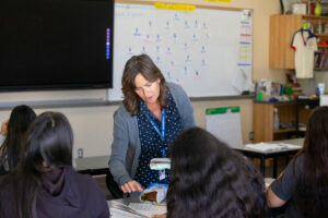 A woman in a gray cardigan and a blue polka dot shirt leans over a student's desk in a classroom.