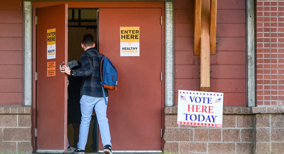 A person wearing a plaid shirt and jeans enters a building with a "vote here today sign" outside.