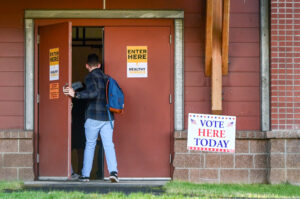 A person wearing a plaid shirt and jeans enters a building with a "vote here today sign" outside.