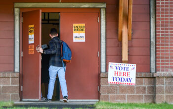 A person wearing a plaid shirt and jeans enters a building with a "vote here today sign" outside.