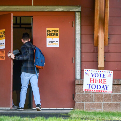 A person wearing a plaid shirt and jeans enters a building with a "vote here today sign" outside.