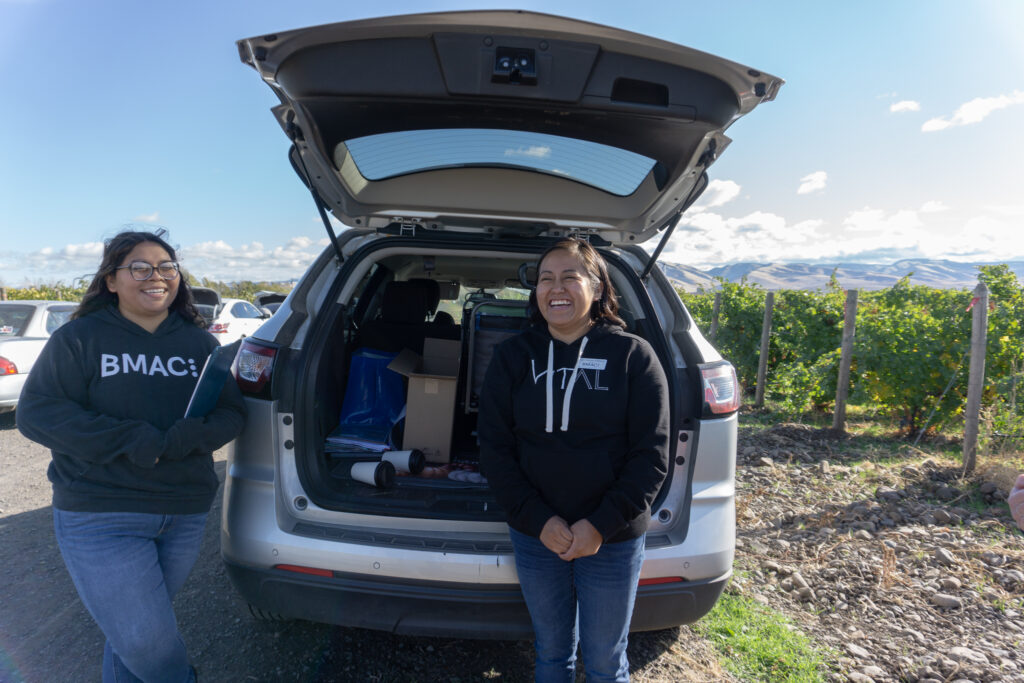 Two smiling women in black hoodies stand in front of a car in a vineyard.