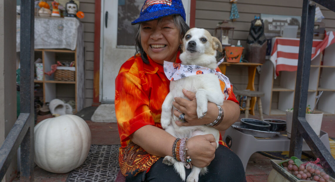 A woman in a red shirt sits on her porch and holds a small dog.